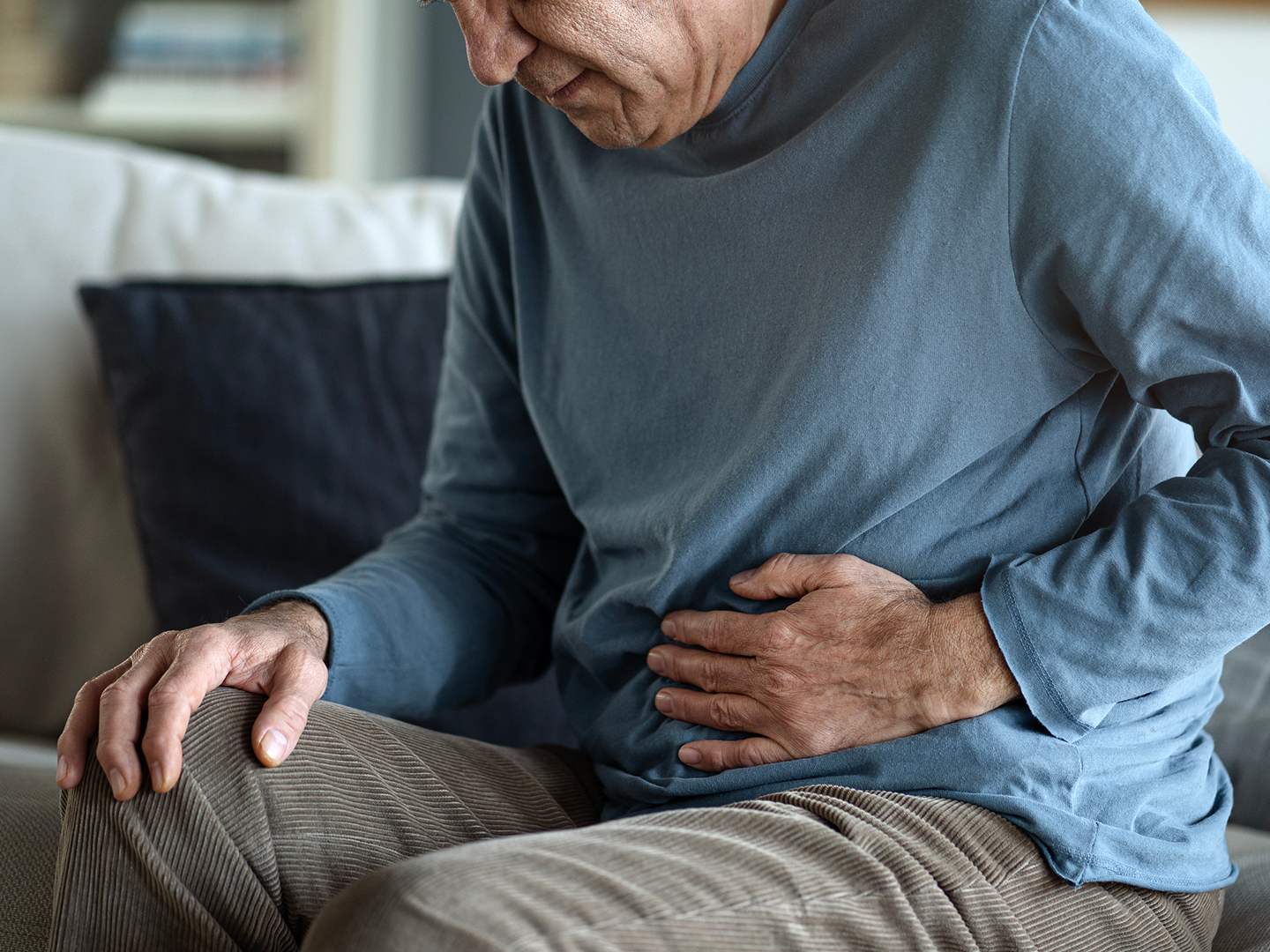 Älterer Mann im blauen Pullover sitzt auf einem Sofa und hält sich mit einer Hand den Bauch.