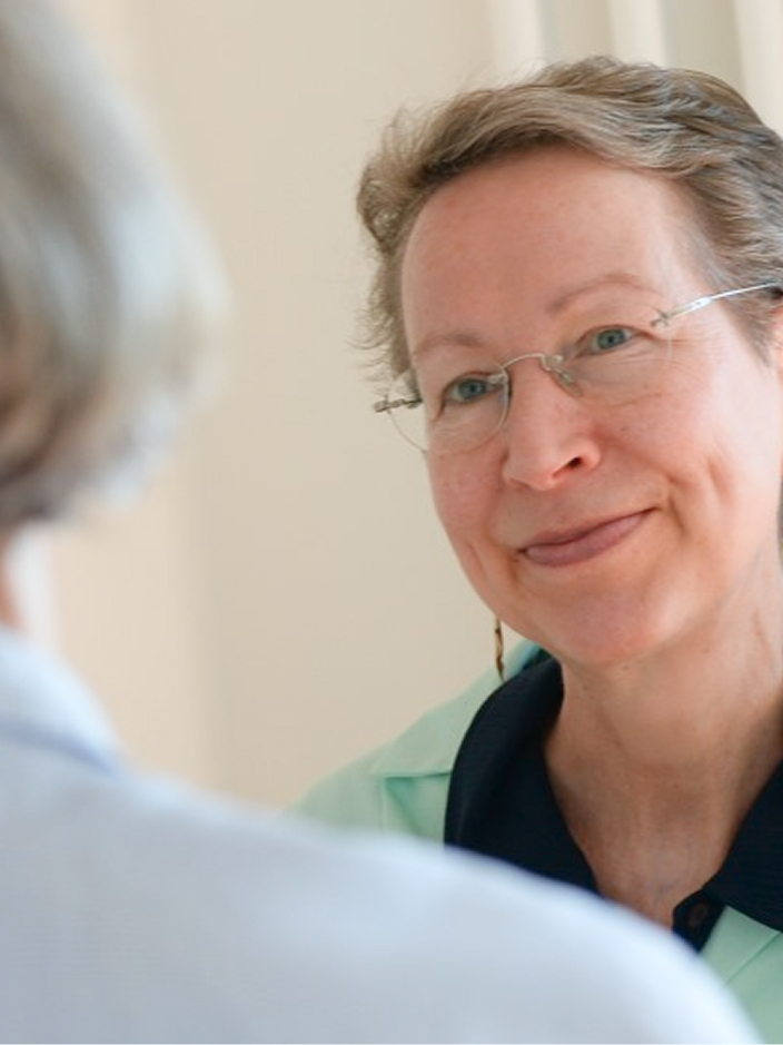 Zwei Frauen in einem Gespräch, eine von hinten sichtbar, die andere lächelt und trägt eine Brille.