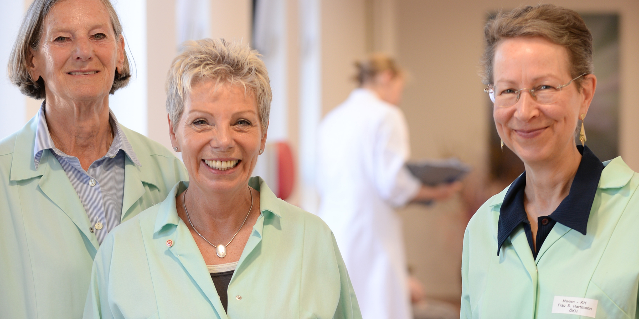 Drei lächelnde, ältere Frauen in grünen Kitteln stehen zusammen im Klinikflur und lächeln in die Kamera. Im Hintergrund arbeitet eine Ärztin im weißen Kittel.