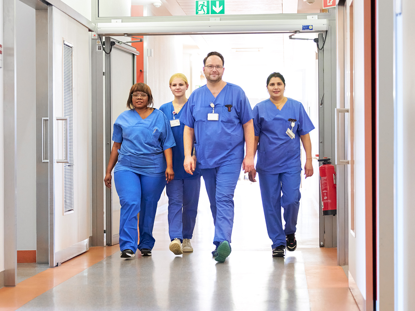 Vier medizinische Fachkräfte in blauer Arbeitskleidung gehen gemeinsam einen hellen Krankenhausflur entlang.