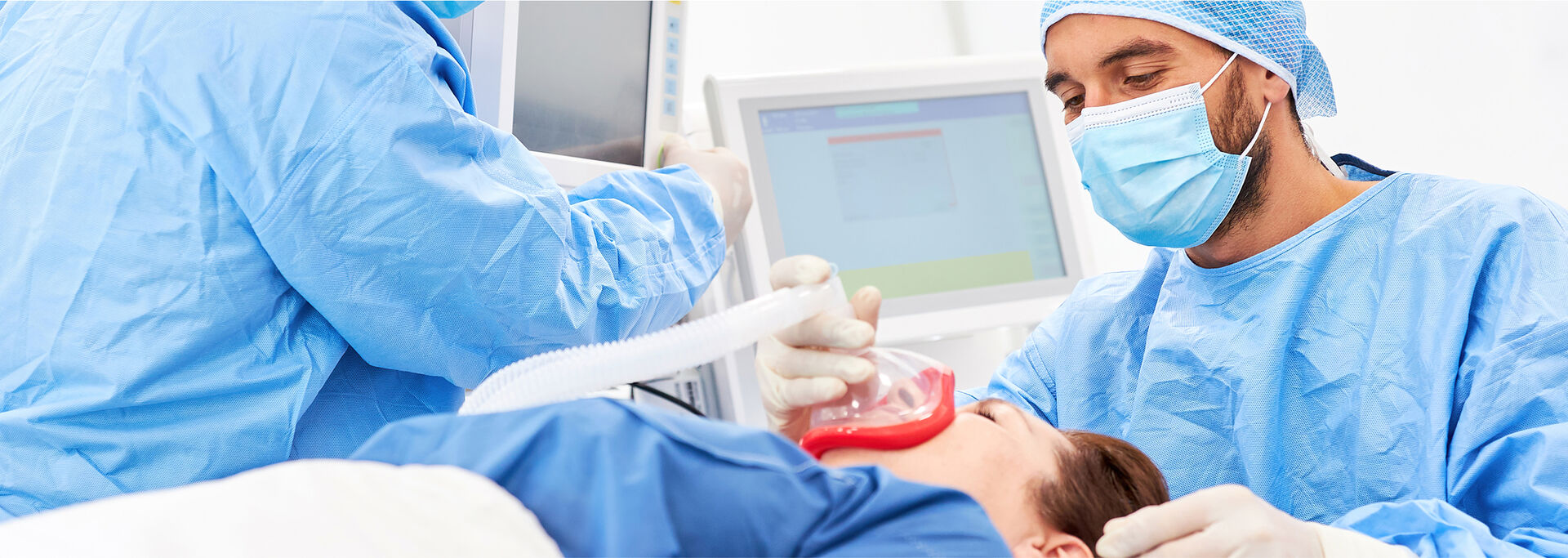 Zwei medizinische Fachkräfte in blauer Schutzkleidung und Masken behandeln einen Patienten, der auf einem Behandlungstisch liegt.