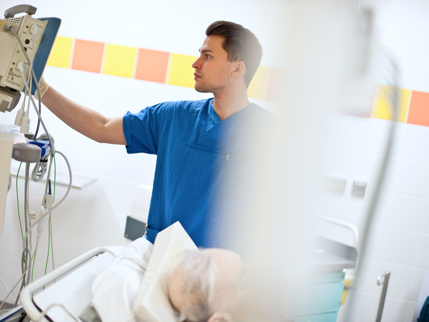 Ein Pfleger in blauer Kleidung steht neben einem Bett und schaut auf ein Tablet, während ein Patient vor ihm im Bett liegt.
