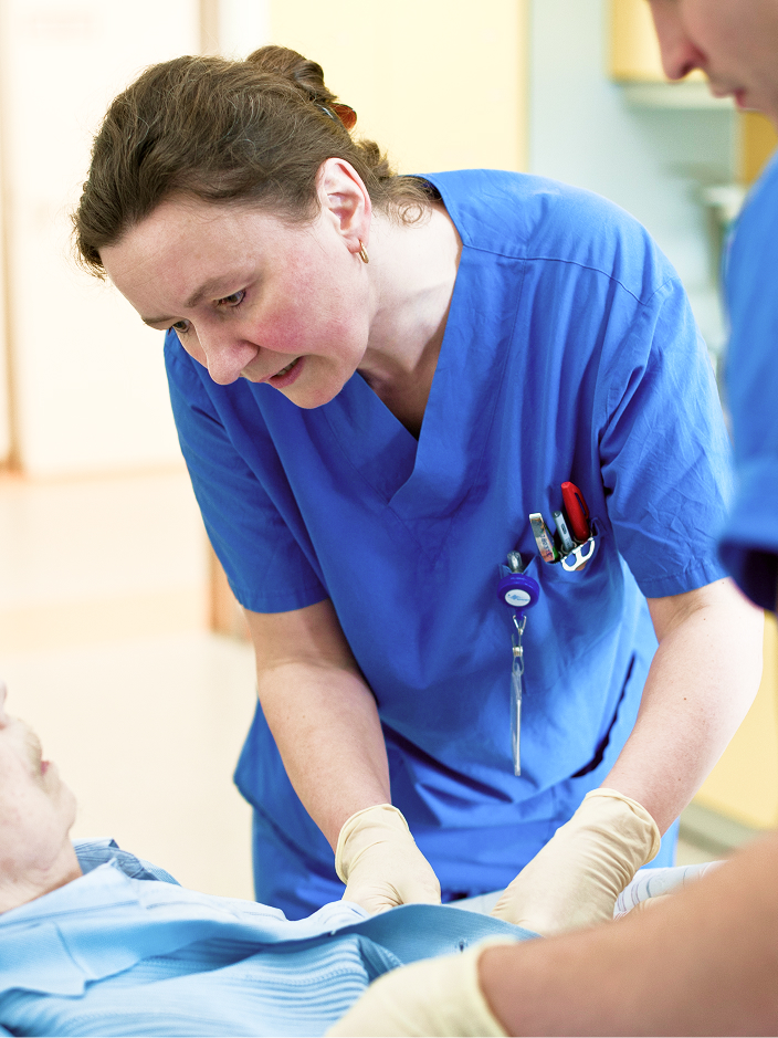 Eine Krankenschwester in blauer Arbeitskleidung kümmert sich um einen Patienten im Bett, während ein Kollege assistiert.