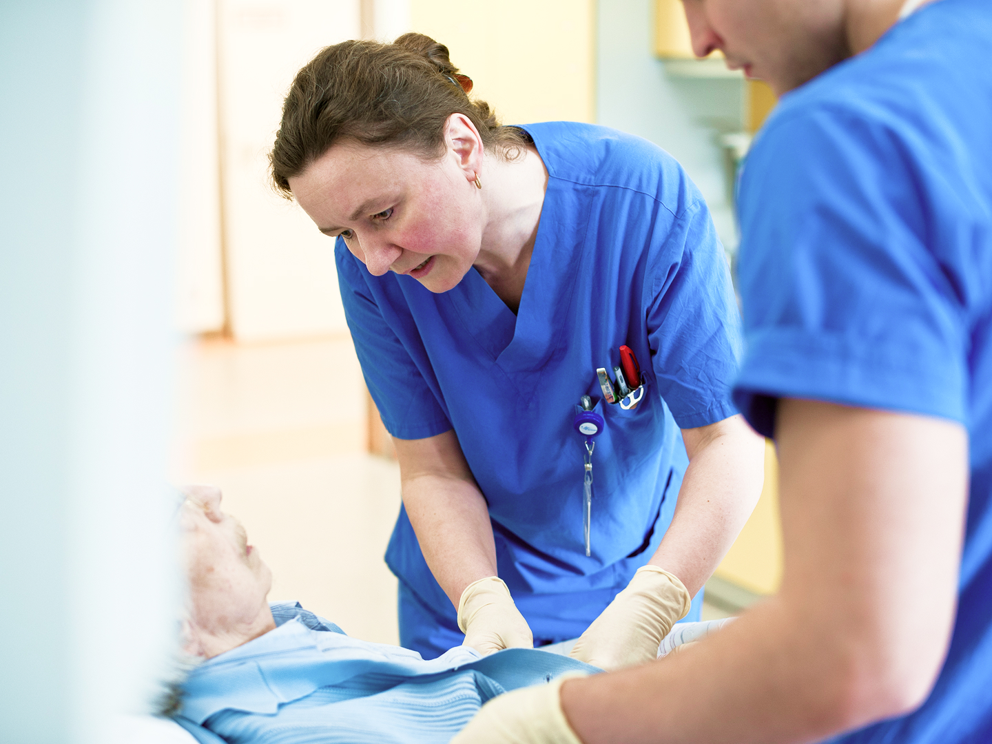 Zwei Pflegekräfte in blauer Arbeitskleidung kümmern sich um einen älteren Patienten im Bett.