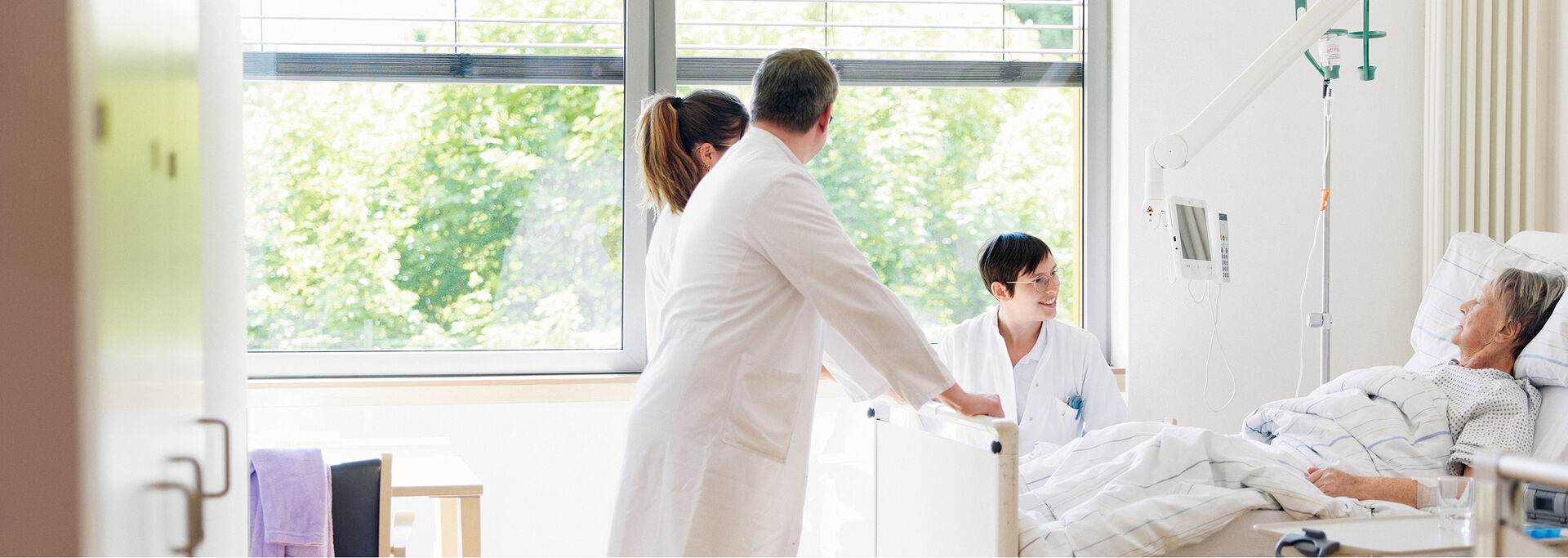 Ärzte stehen in einem hellen Krankenhauszimmer am Bett eines Patienten. Tageslicht strömt durch das Fenster.