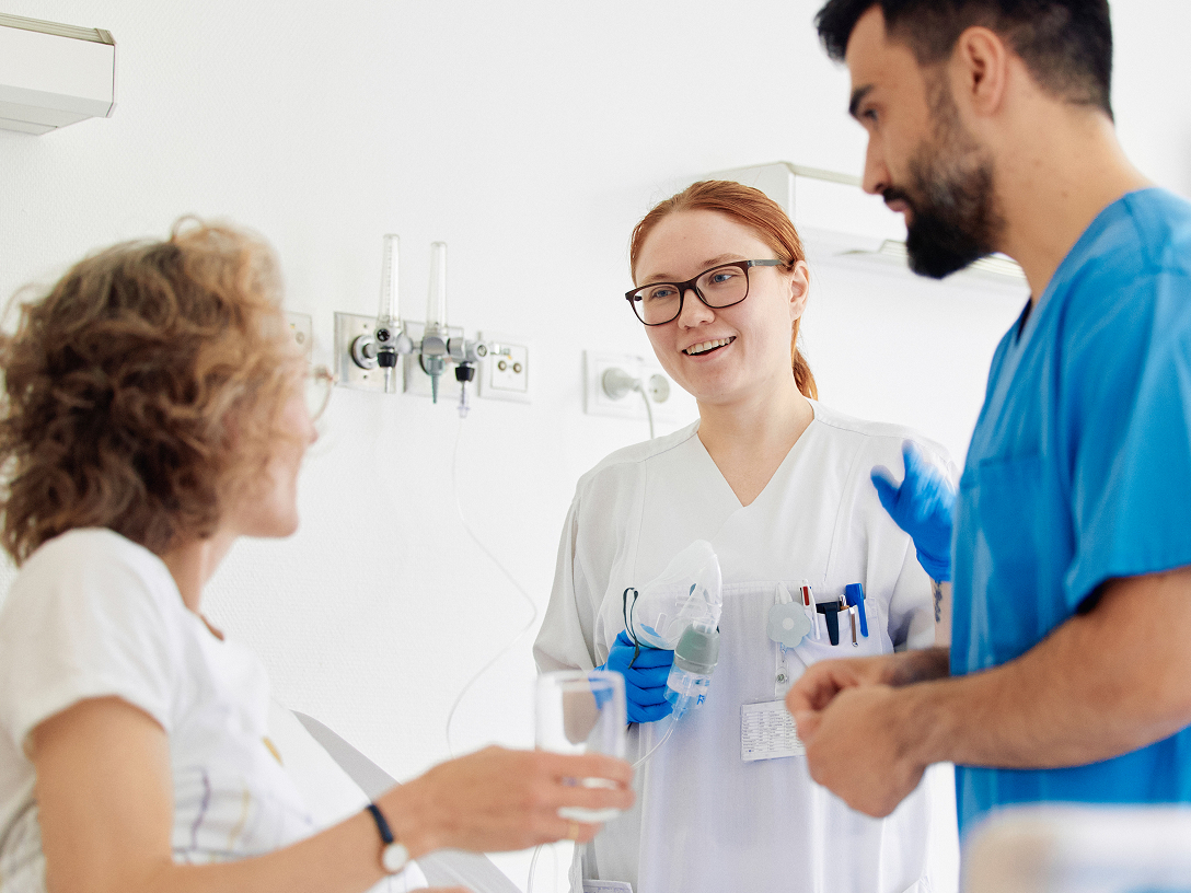 Zwei medizinische Fachkräfte stehen am Bett einer Patientin, die ein Glas Wasser in der Hand hält.