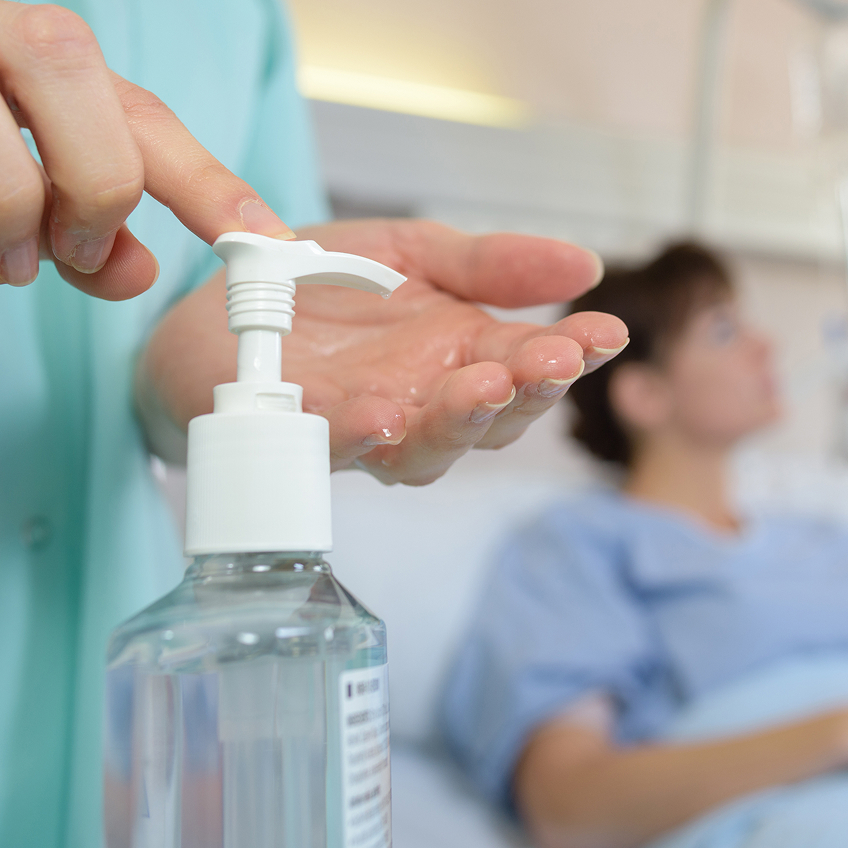 Eine medizinische Fachkraft betätigt einen Spender mit Desinfektionsmittel, während im Hintergrund eine Patientin im Bett liegt.