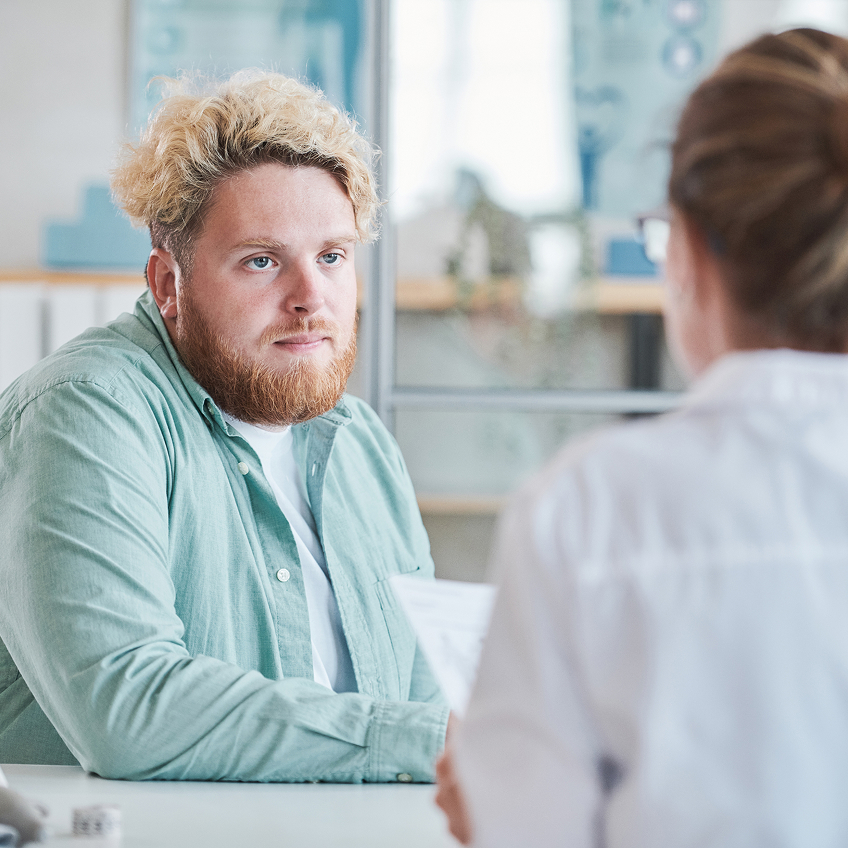 Patient mit blonden Locken und Bart sitzt an einem Tisch und hört einer Ärztin zu, die ihm gegenübersitzt.
