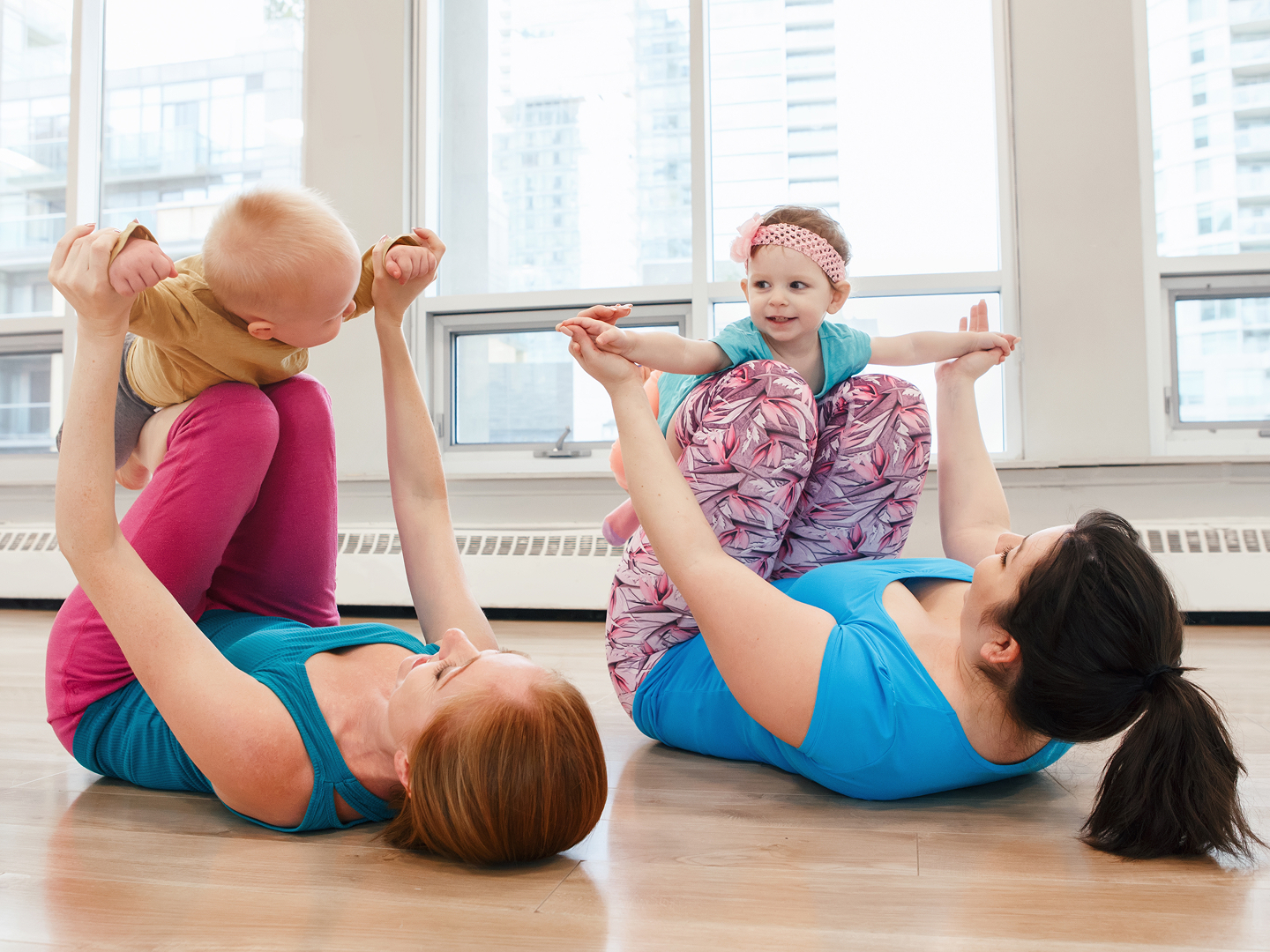 Zwei Frauen machen Yoga-Übungen auf dem Rücken, während sie jeweils ein Kleinkind auf ihren Knien halten. Helle Fenster im Hintergrund.