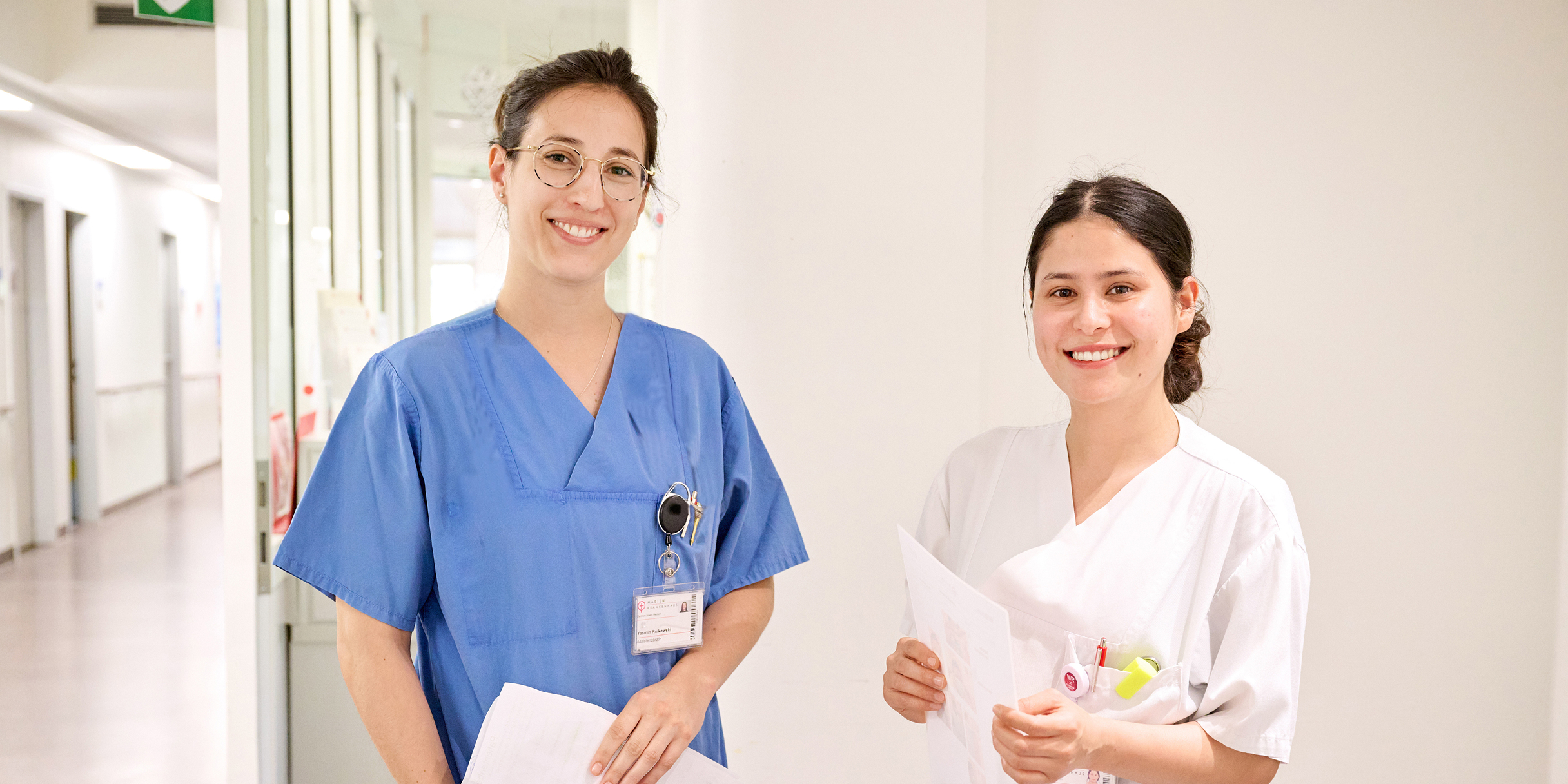 Zwei lächelnde Pflegekräfte stehen in einem Krankenhausflur und halten Dokumente in der Hand.