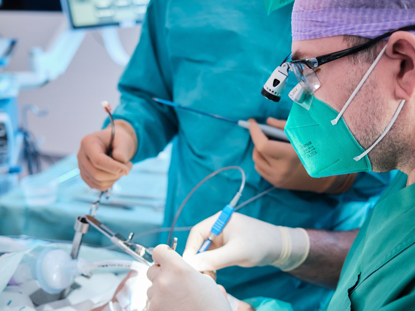 Zwei Chirurgen in OP-Kleidung führen eine Operation durch, einer hält ein chirurgisches Instrument, der andere assistiert.