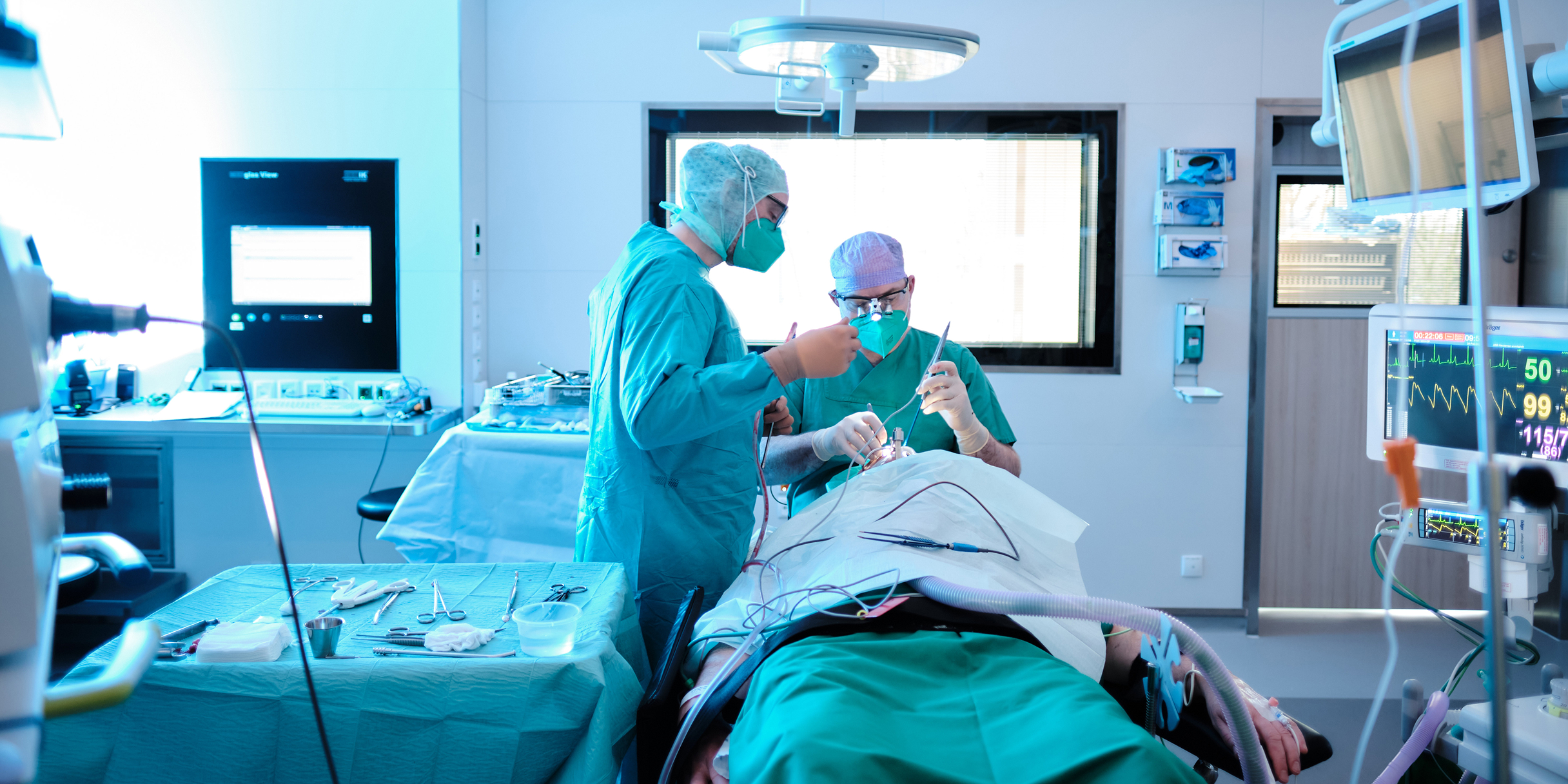 Zwei Chirurgen in OP-Kleidung führen eine Operation an einem Patienten durch, umgeben von medizinischen Geräten.