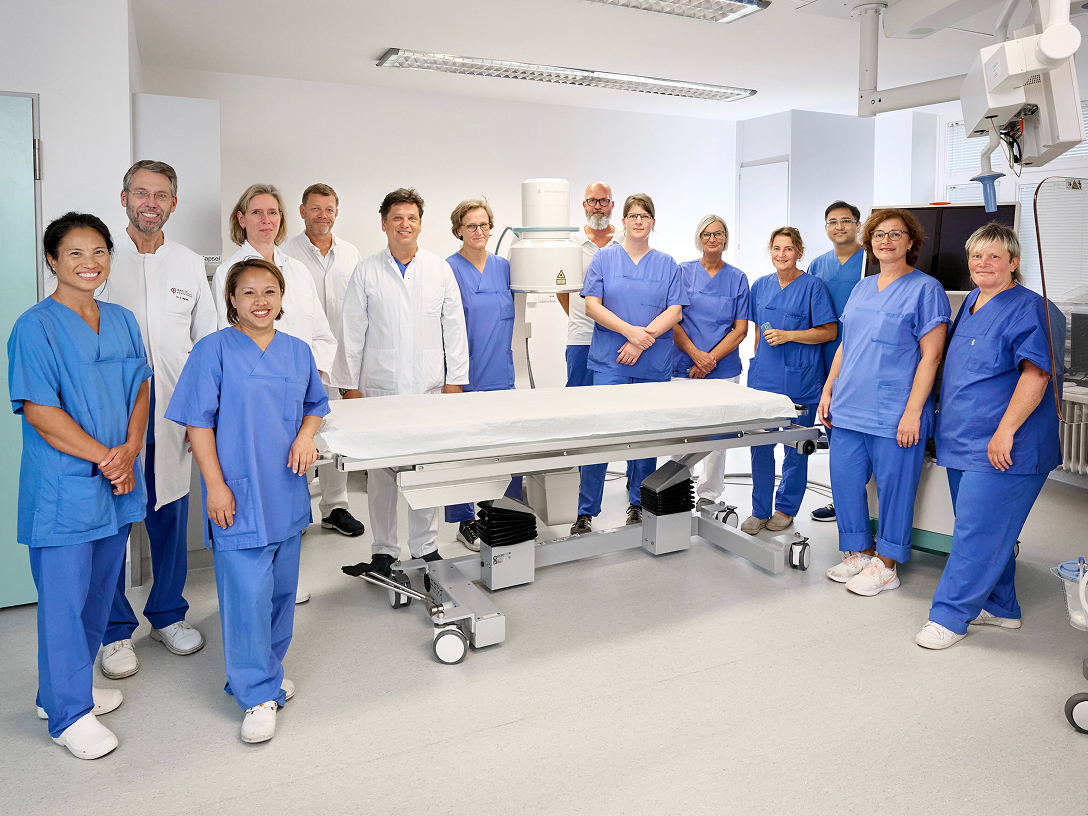 Gruppe von medizinischem Personal in blauer und weißer Arbeitskleidung steht in einem modernen Raum um einen Behandlungstisch herum. Alle schauen zur Kamera.
