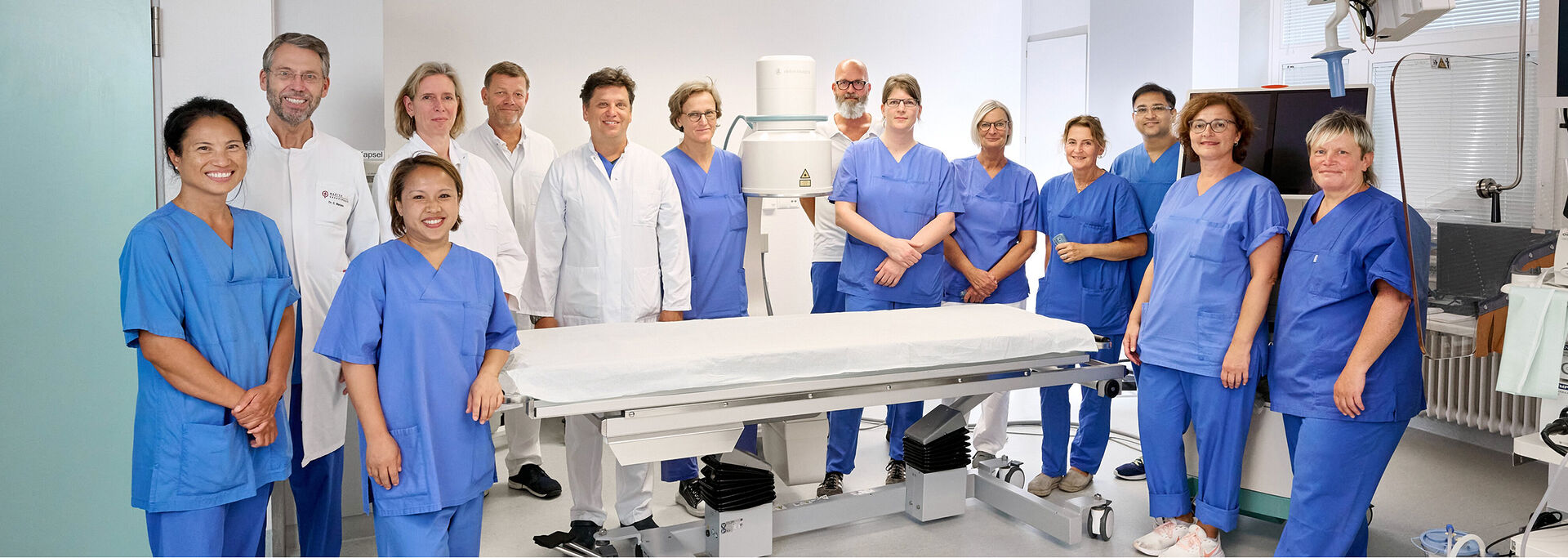 Gruppe von medizinischem Personal in blauer und weißer Arbeitskleidung steht in einem modernen Raum um einen Behandlungstisch herum. Alle schauen zur Kamera.