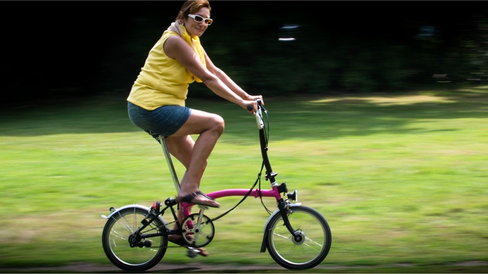 Frau in gelbem Oberteil fährt auf einem pinken Fahrrad über eine grüne Wiese.