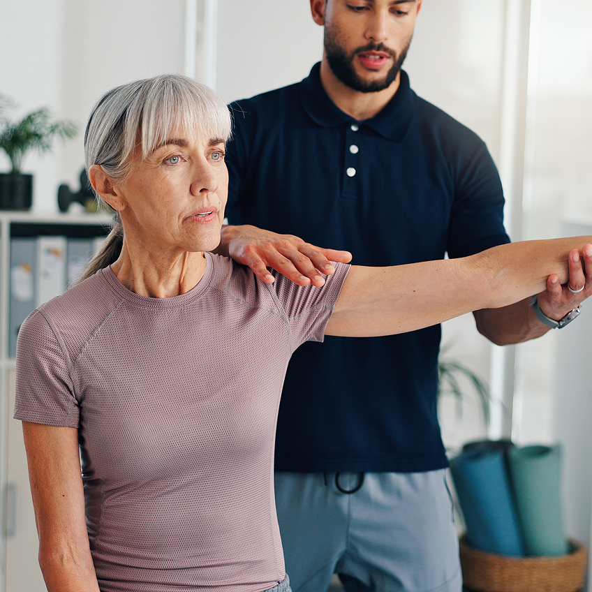 Ein junger Mann hilft einer älteren Frau, ihren Arm zu strecken, beide stehen in einem Trainingsraum.