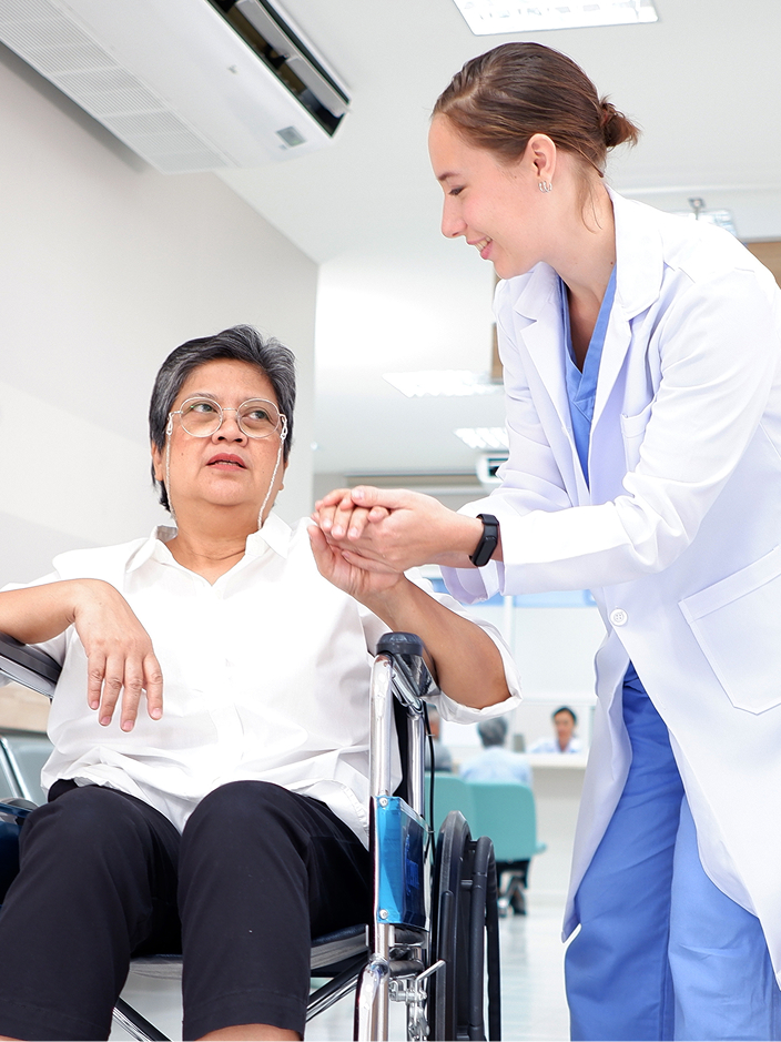 Ärztin hilft einer Patientin mit Brille und Rollstuhl in einem modernen Klinikflur.