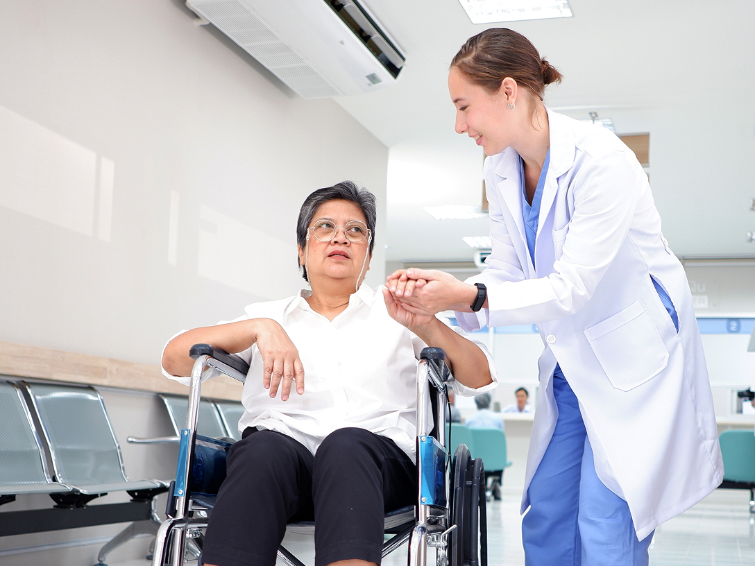 Ärztin hilft einer Patientin mit Brille und Rollstuhl in einem modernen Klinikflur.