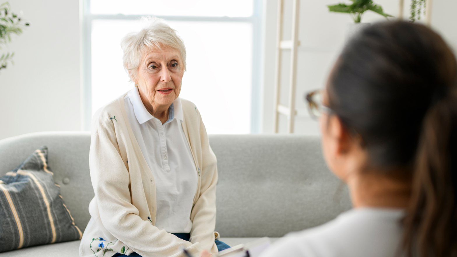 Eine ältere Patientin in heller Kleidung sitzt auf einem grauen Sofa und spricht mit einer Ärztin, die ihr gegenübersitzt.