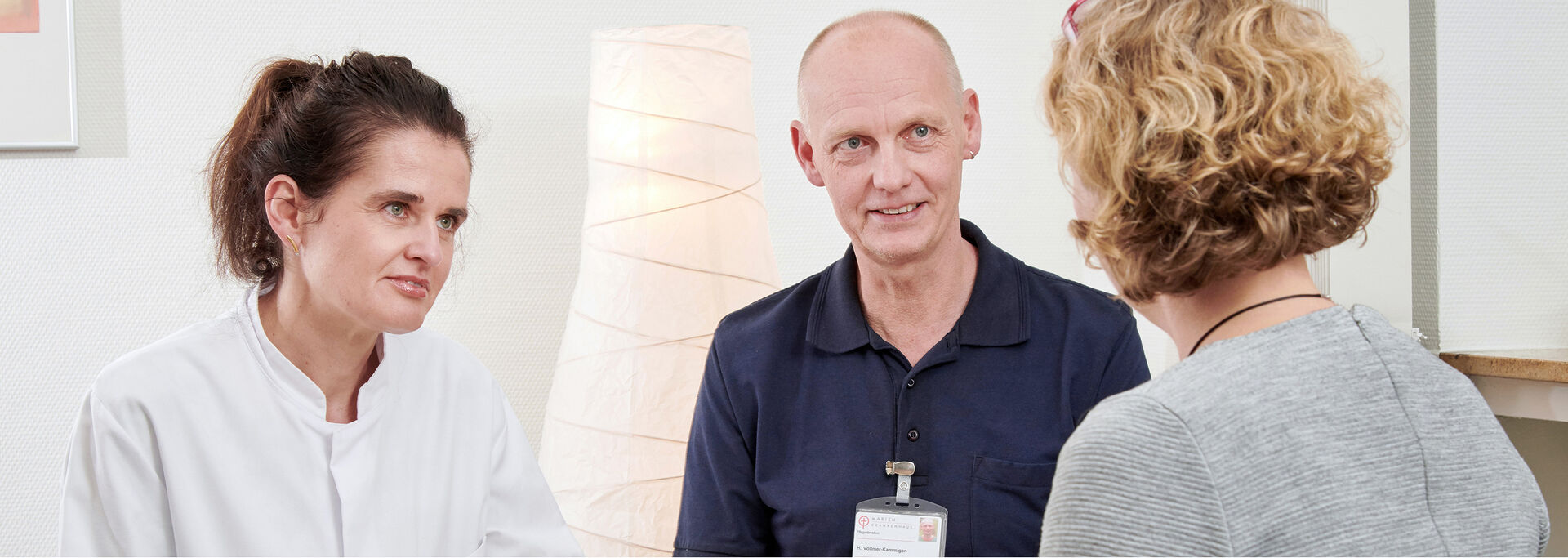 Drei Personen in einem Gespräch, eine in weißer Kleidung, eine in dunkler Kleidung und eine mit lockigem, blondem Haar.