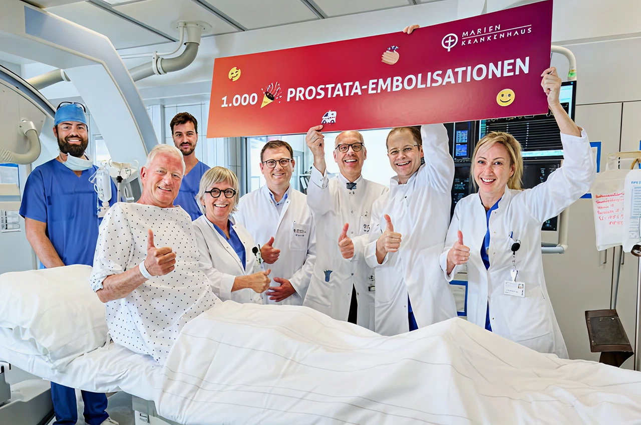 Gruppe von Ärzten steht am Bett eines Patienten, sie halten ein Banner mit der Aufschrift "1.000 PROSTATA-EMBOLISATIONEN" nach oben und freuen sich.
