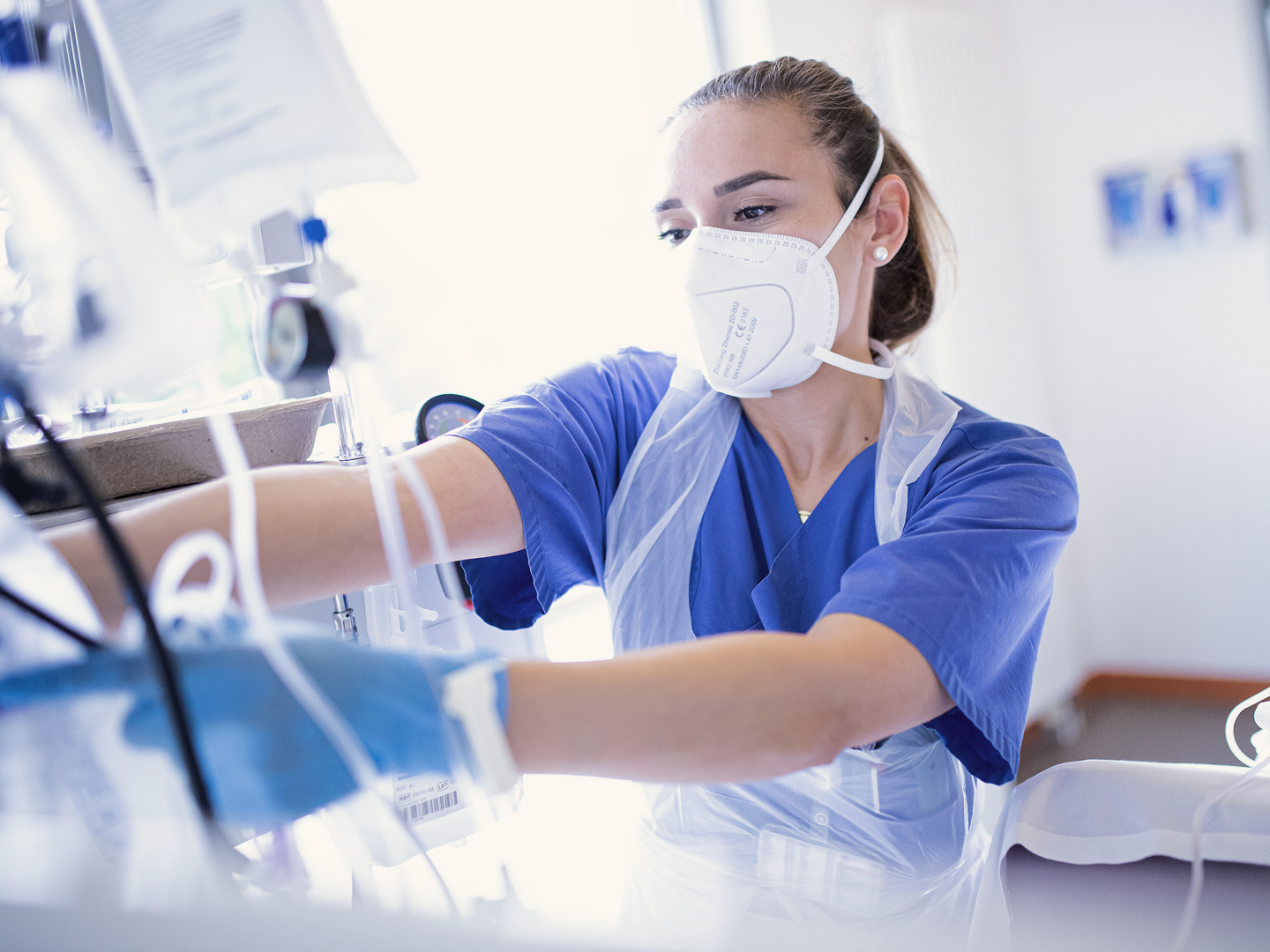 Eine medizinische Fachkraft mit blauer Arbeitskleidung und Maske bedient medizinische Geräte in einem klinischen Umfeld.