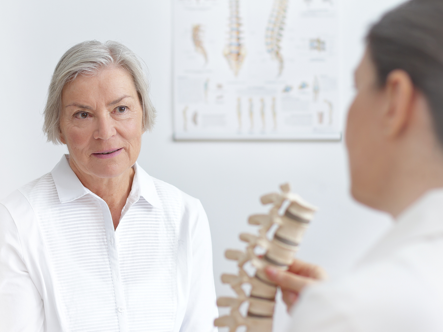 Ältere Frau hört einer Ärztin zu, die ihr in heller, moderner Umgebung ein Modell der Wirbelsäule zeigt.