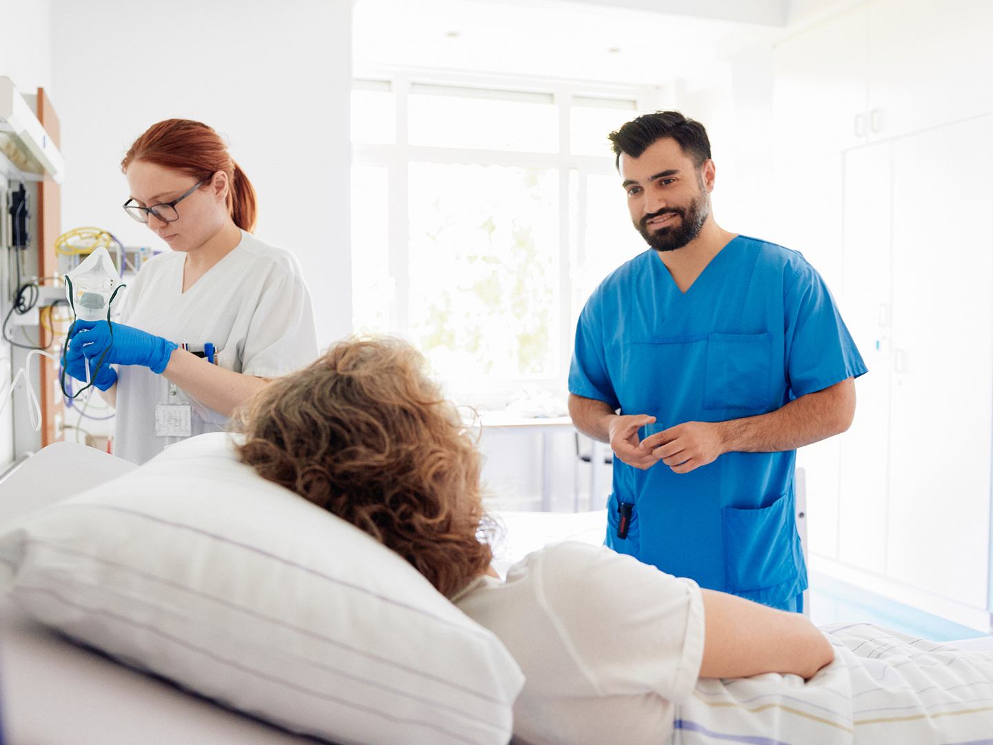 Zwei medizinische Fachkräfte kümmern sich um eine Patientin, die in einem hellen Krankenhauszimmer im Bett liegt.