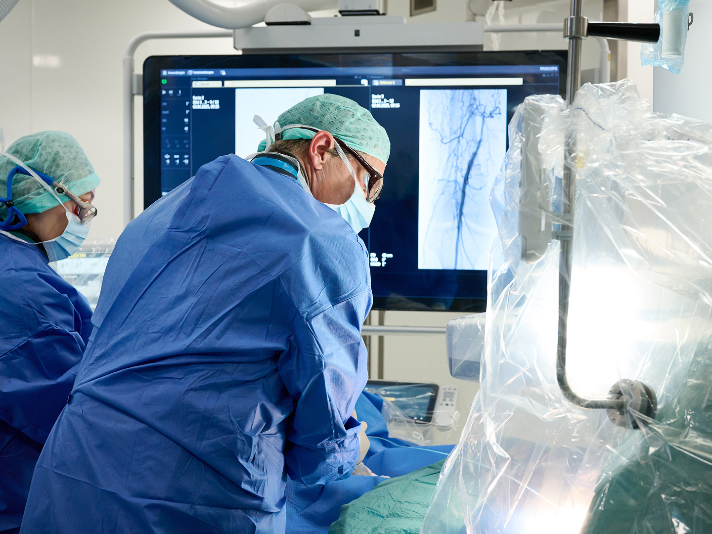 Zwei Chirurgen mit blauen OP-Kitteln und Hauben arbeiten an einem Patienten, während ein Monitor medizinische Bilder zeigt.