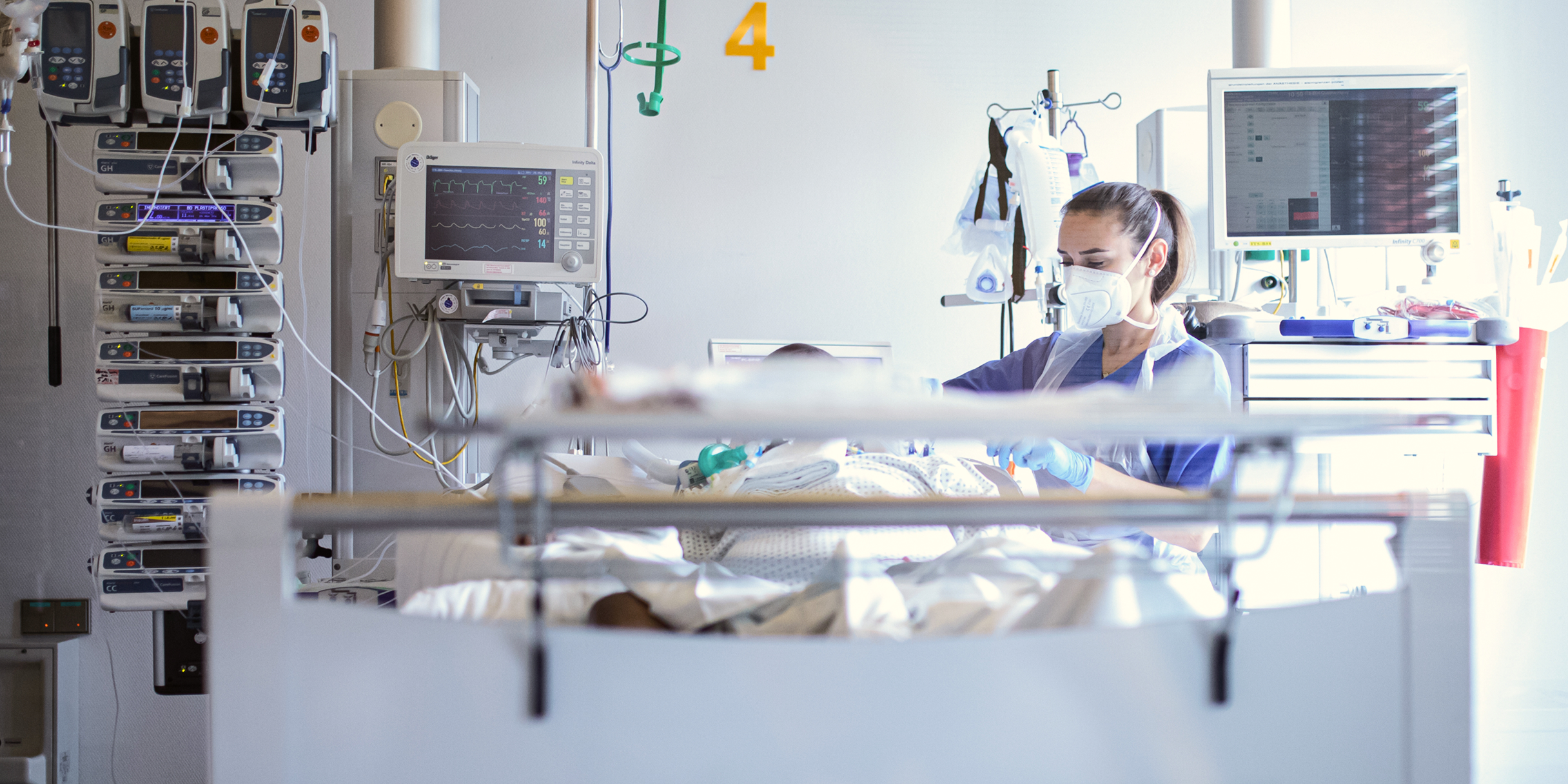 Krankenhauszimmer mit einer medizinischer Fachkraft, die sich um einen Patienten kümmert, umgeben von Geräten.