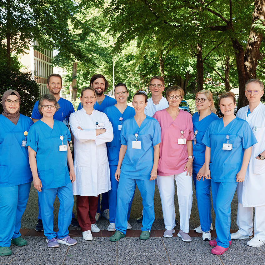 Gruppe von medizinischen Fachkräften in Arbeitskleidung, lächelnd im Freien vor Bäumen.