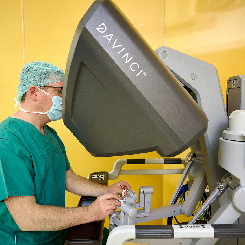 Ein Arzt in grüner OP-Kleidung bedient ein Da Vinci-Operationssystem in einem gelben Raum.