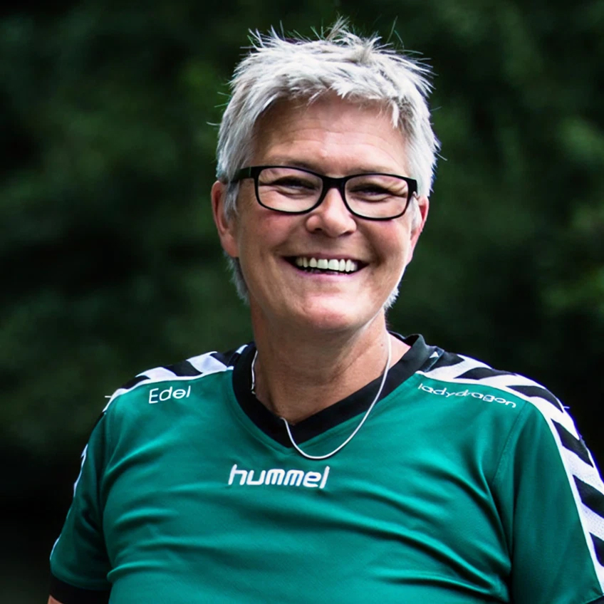 Lächelnde Person mit kurzen grauen Haaren, Brille und grünem Sporttrikot, vor unscharfem Hintergrund.