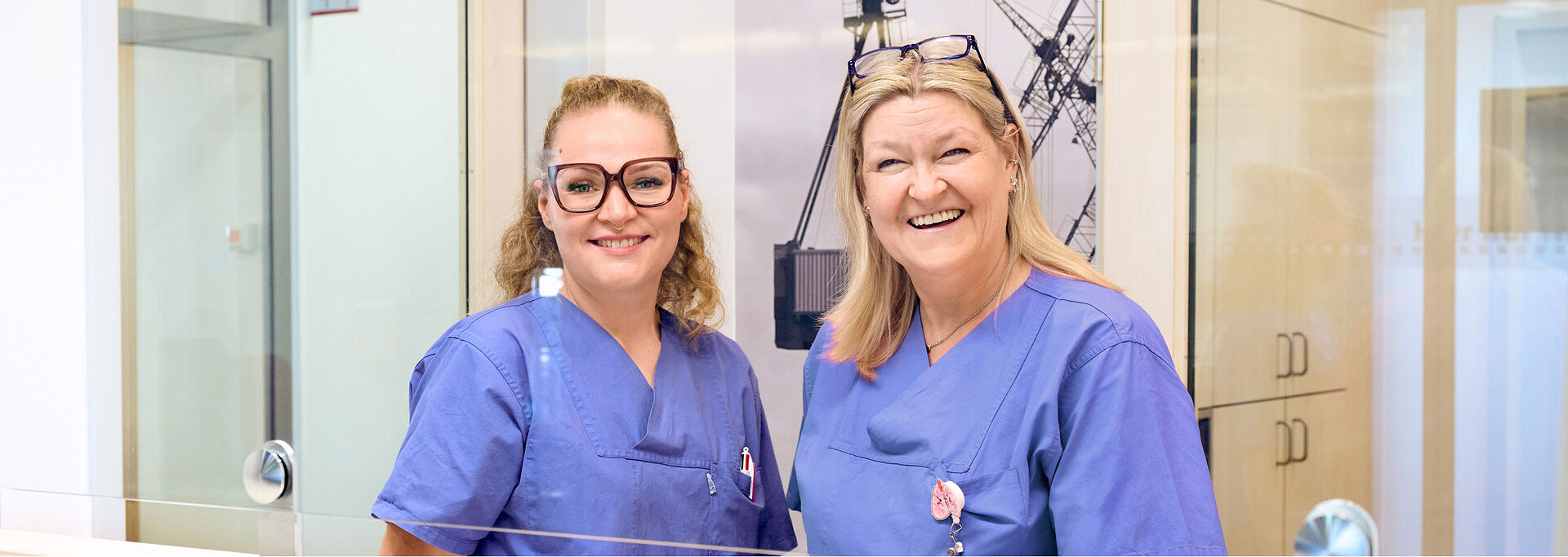 Zwei lächelnde, blonde Frauen in blauer Arbeitskleidung stehen hinter einem Empfangstresen.