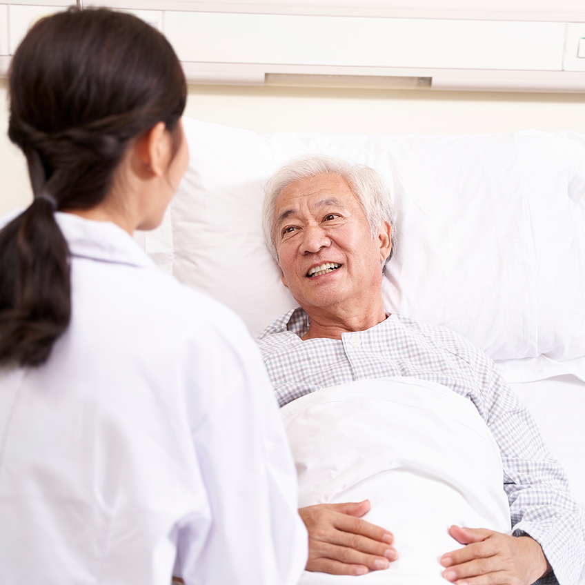 Ein älterer Mann liegt im Krankenhausbett und lächelt eine dunkelhaarige Ärztin an, die vor ihm steht.