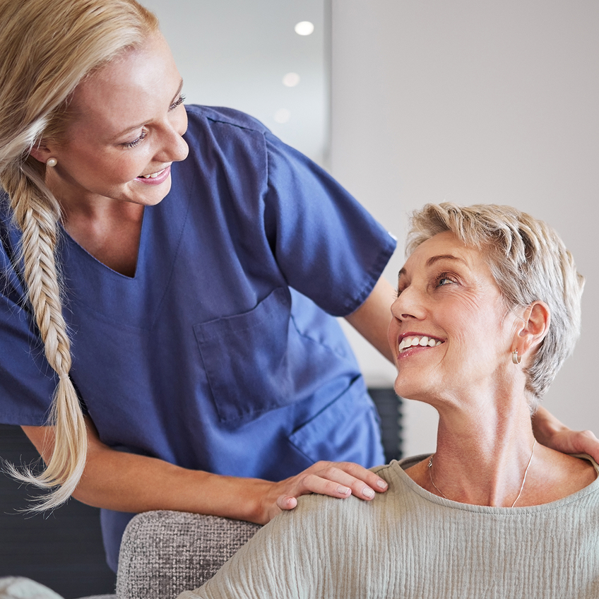 Weibliche Pflegekraft beugt sich von hinten zu einer lächelnden Patientin hinunter und spricht sie freundlich an.