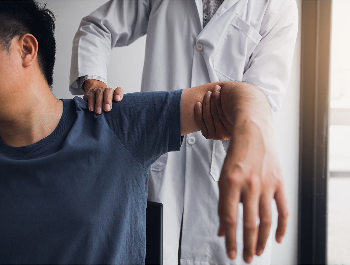 Ein Arzt untersucht einen Patienten, der sich zur Seite dreht, während der Arzt sein Schultergelenk begutachtet.