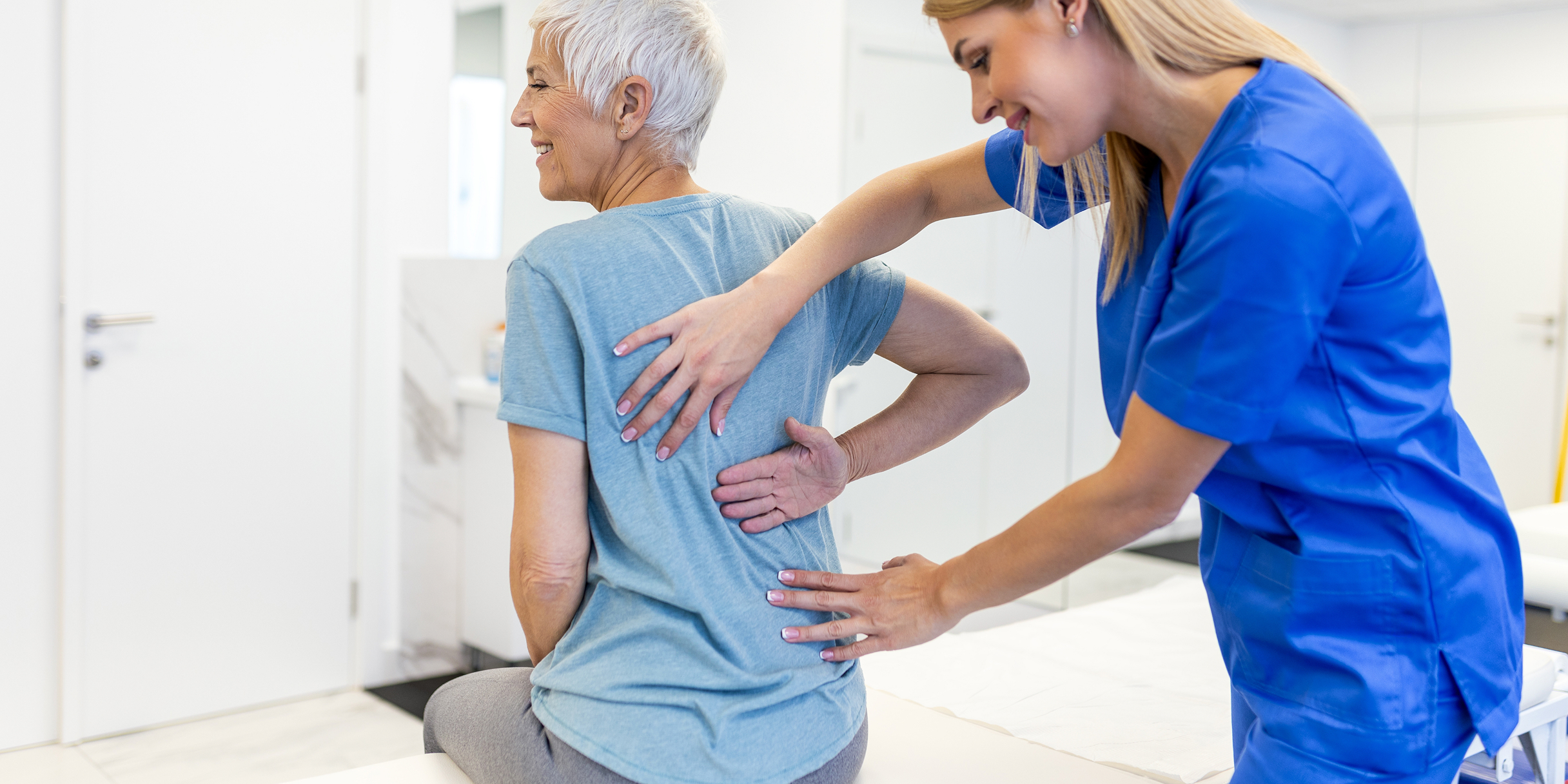 Eine ältere Patientin sitzt auf einer Liege und wird von einer medizinischen Fachkraft in blauer Uniform am Rücken untersucht.