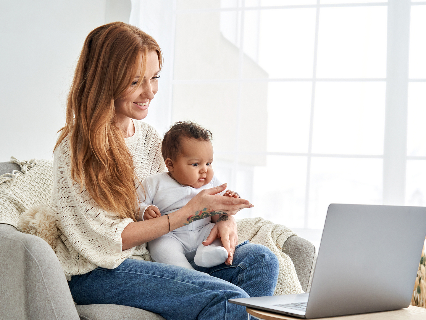 Eine lächelnde Frau sitzt mit einem Baby auf dem Schoß vor einem Laptop in einem hellen Raum.