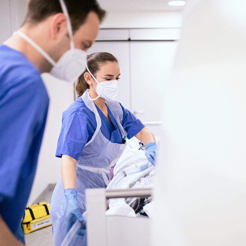 Zwei Personen in blauen Arbeitskleidung und Mundschutz führen einen Schlauch zum Bett eines Patienten.