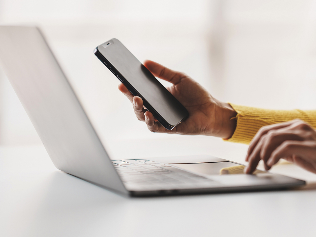 Eine Hand hält ein Smartphone, während die andere Hand auf einer Laptop-Tastatur tippt.