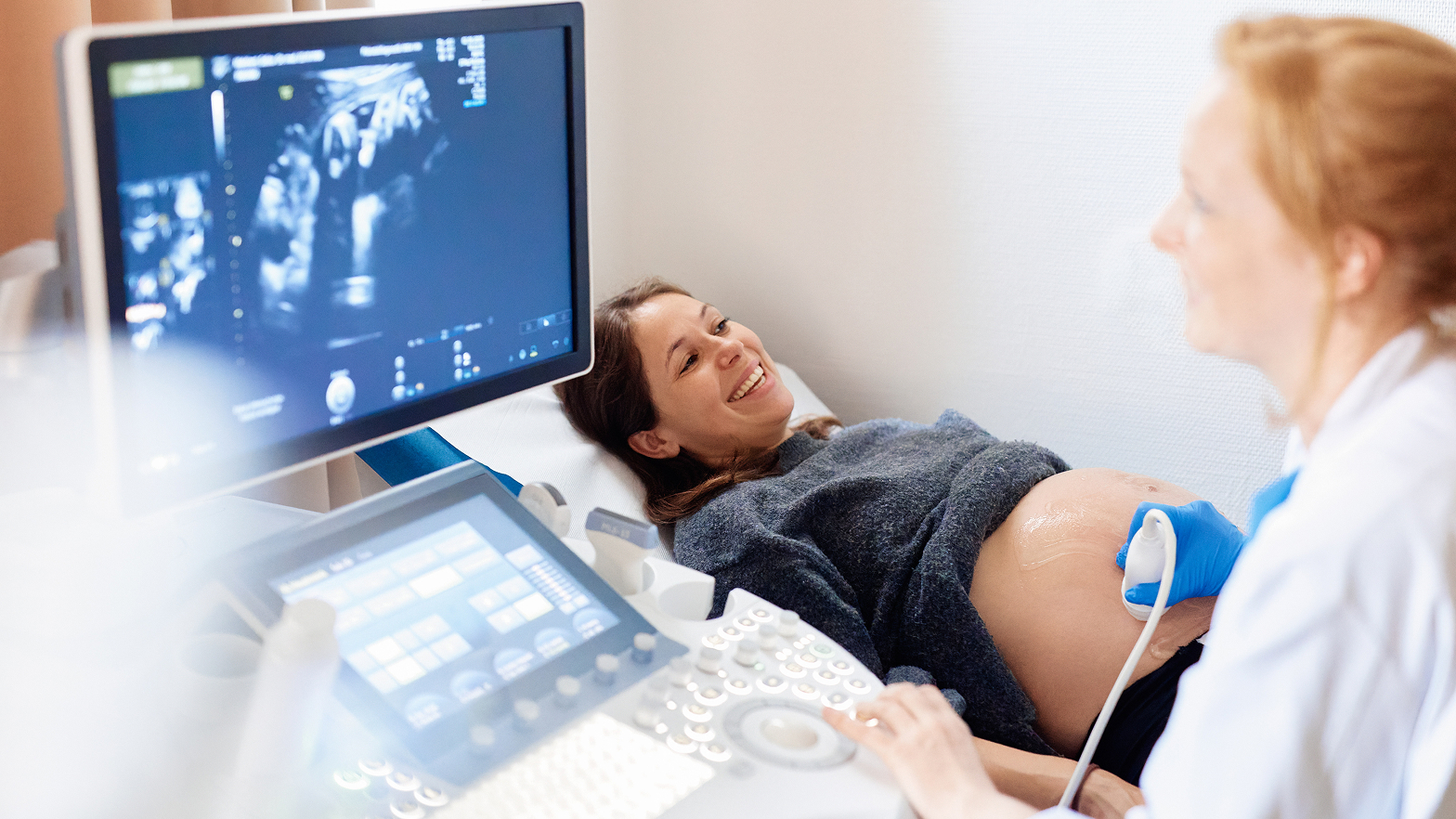 Liegende, schwangere Frau lächelt während einer Ultraschalluntersuchung, eine Ärztin bedient das Gerät und schaut dabei auf einen Monitor.