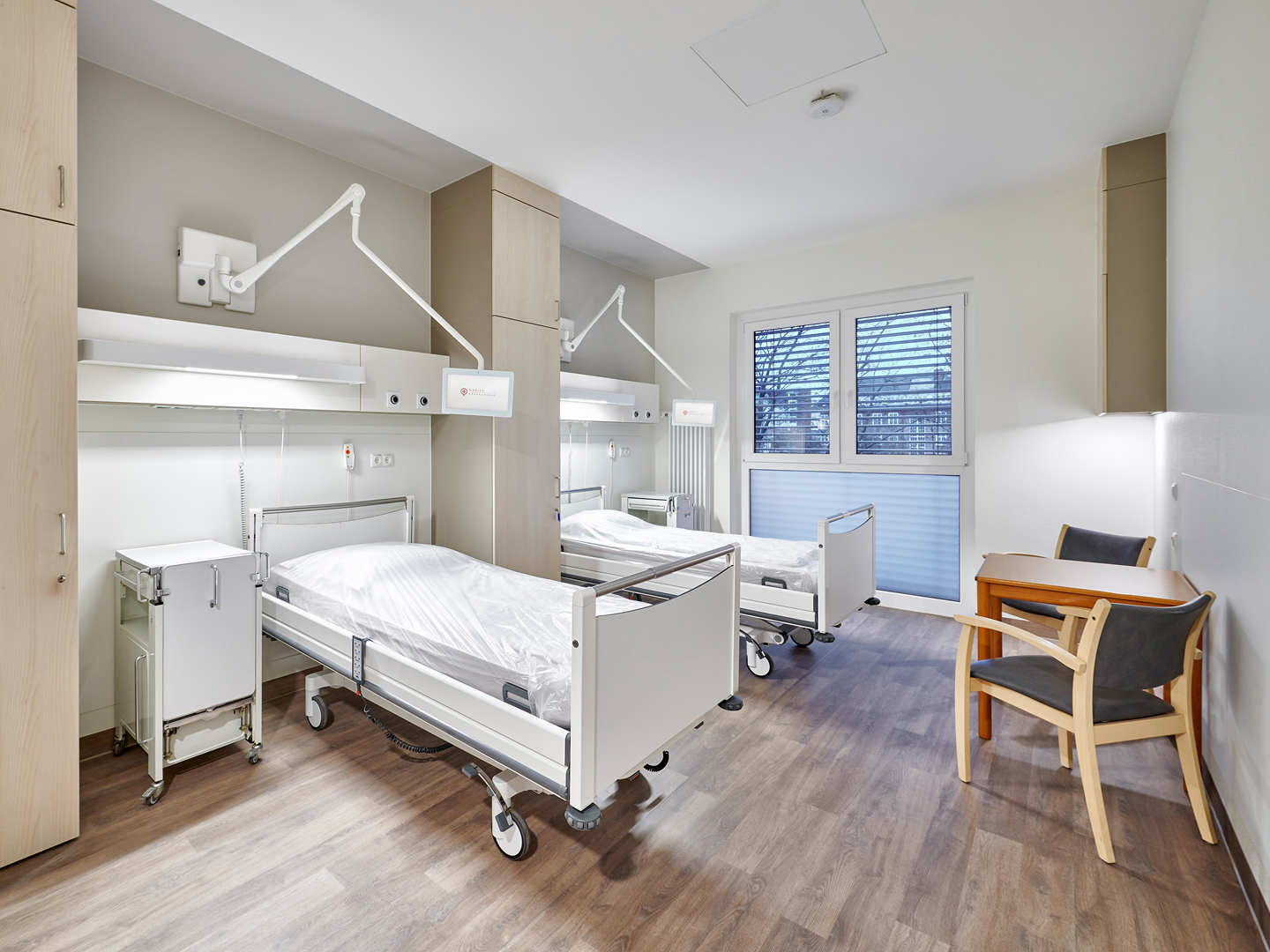 Helles und modern eingerichtetes Krankenhauszimmer mit zwei Betten, Schränken und Sitzgelegenheiten.