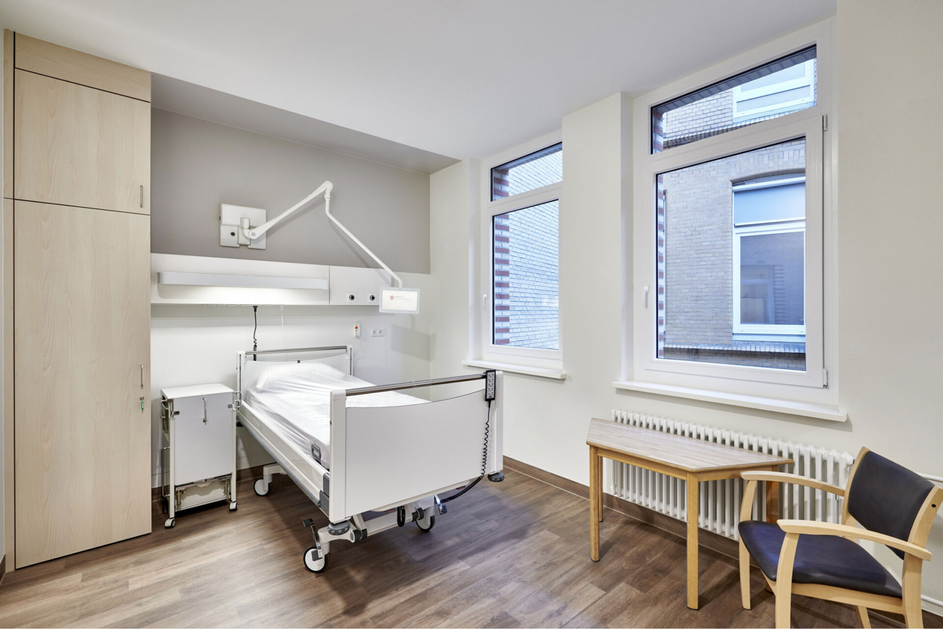 Helles und modern eingerichtetes Krankenhauszimmer mit zwei Betten, Schränken und Sitzgelegenheiten.