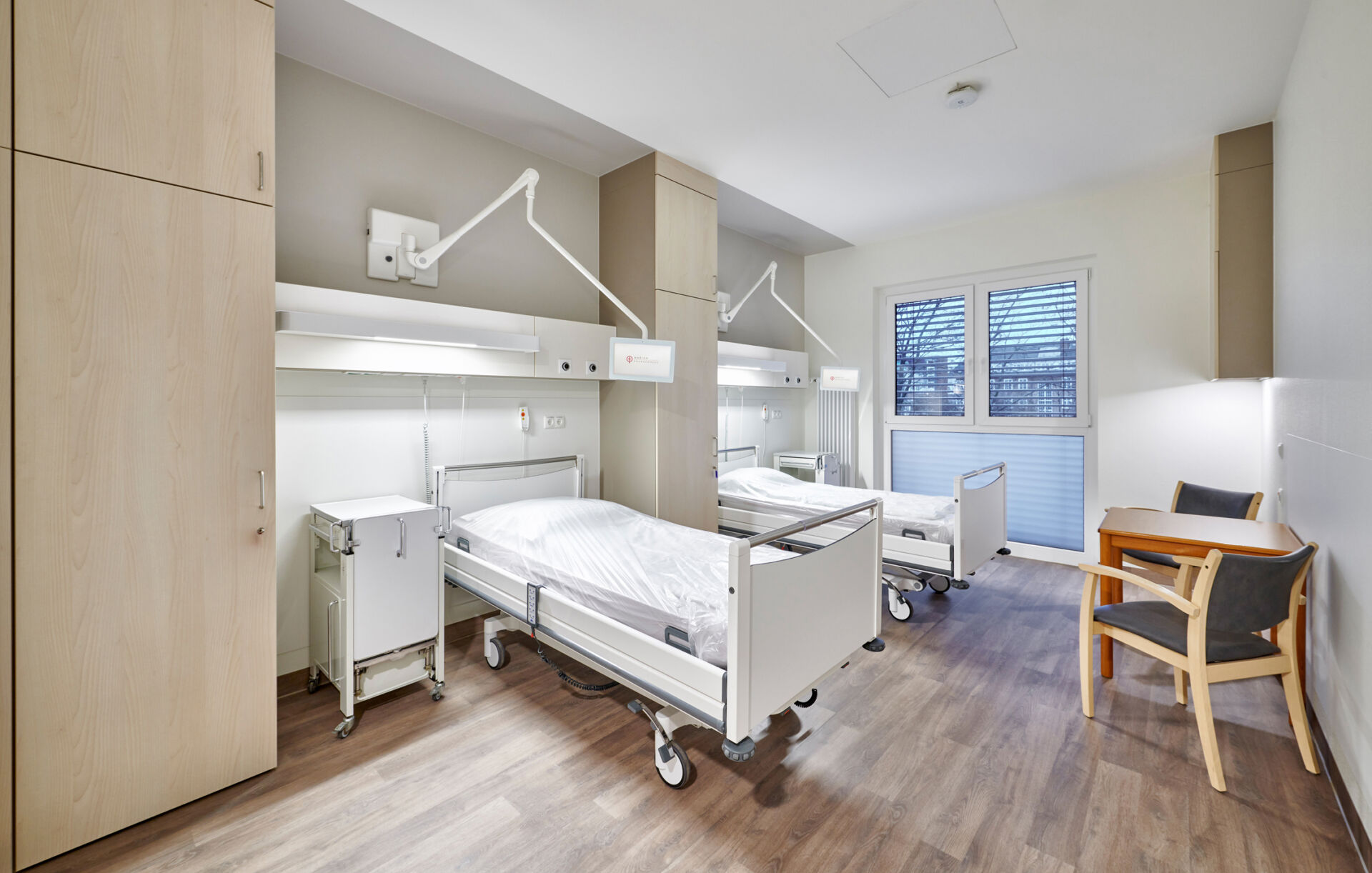 Helles und modern eingerichtetes Krankenhauszimmer mit zwei Betten, Schränken und Sitzgelegenheiten.