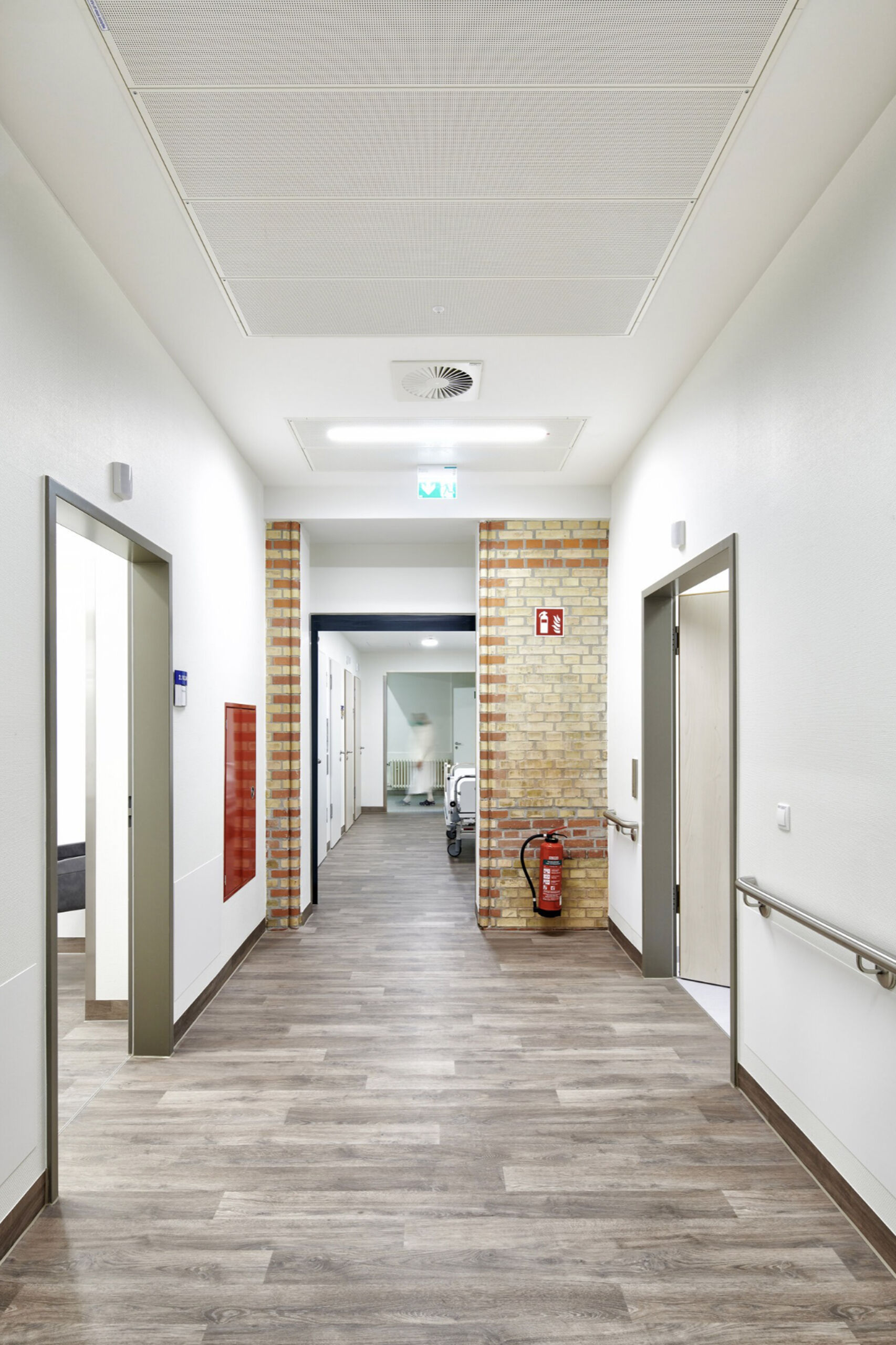Heller Klinikflur mit Türen auf beiden Seiten, Holzfußboden und einem Feuerlöscher an der Wand.