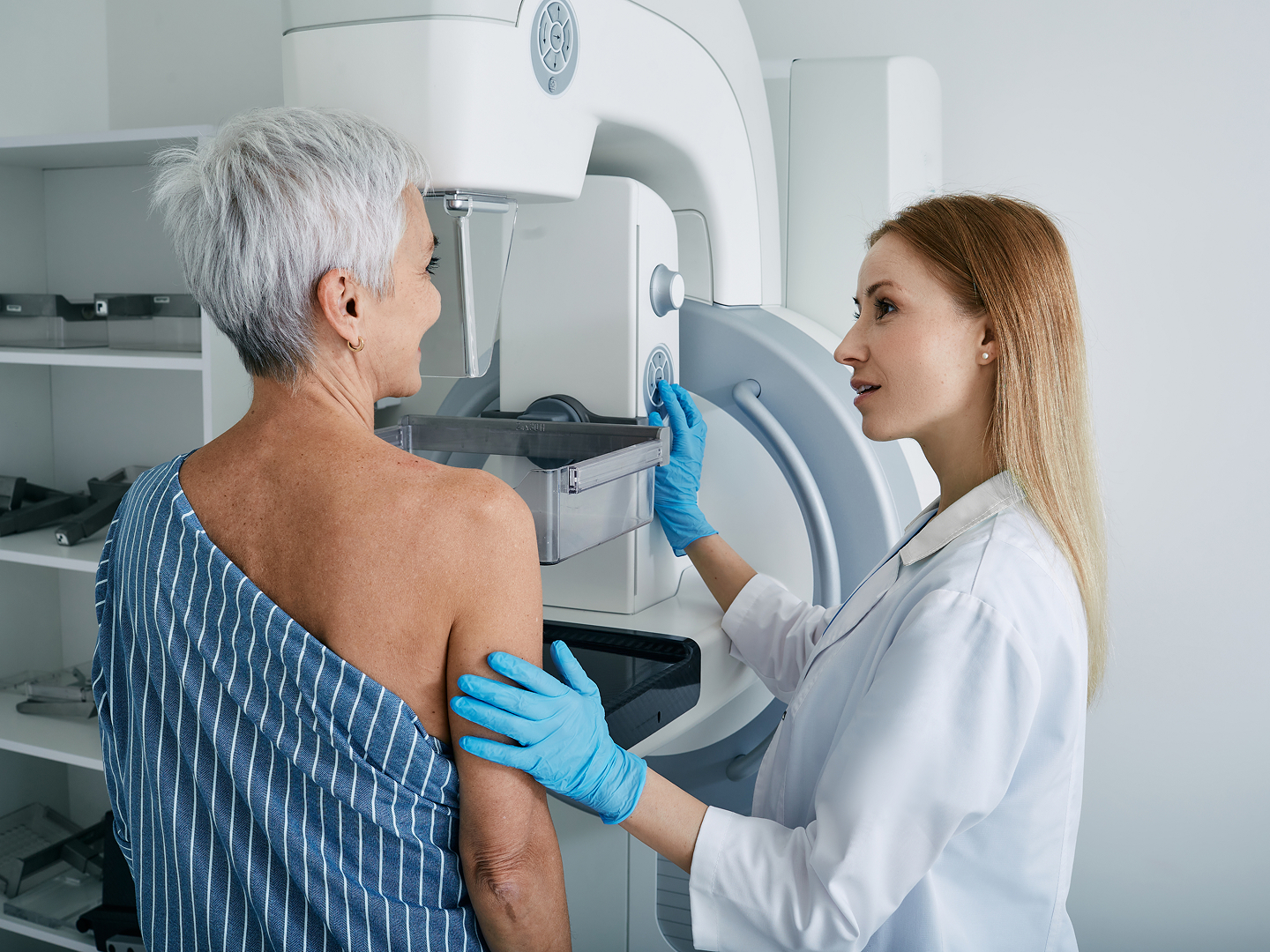 Eine Frau mit kurzen, grauen Haaren steht vor einem Mammografie-Gerät, während eine Ärztin sie an der Schulter berührt und etwas erklärt.