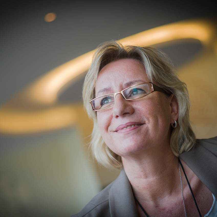 Frau mit blonden Haaren und Brille lächelt in einem modernen Innenraum mit sanfter Beleuchtung.