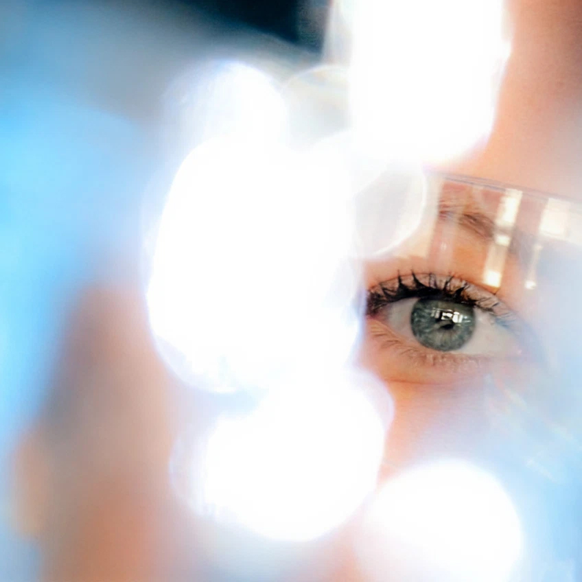 Nahaufnahme eines Auges hinter einer Schutzbrille, umgeben von unscharfen, leuchtenden Lichtern.