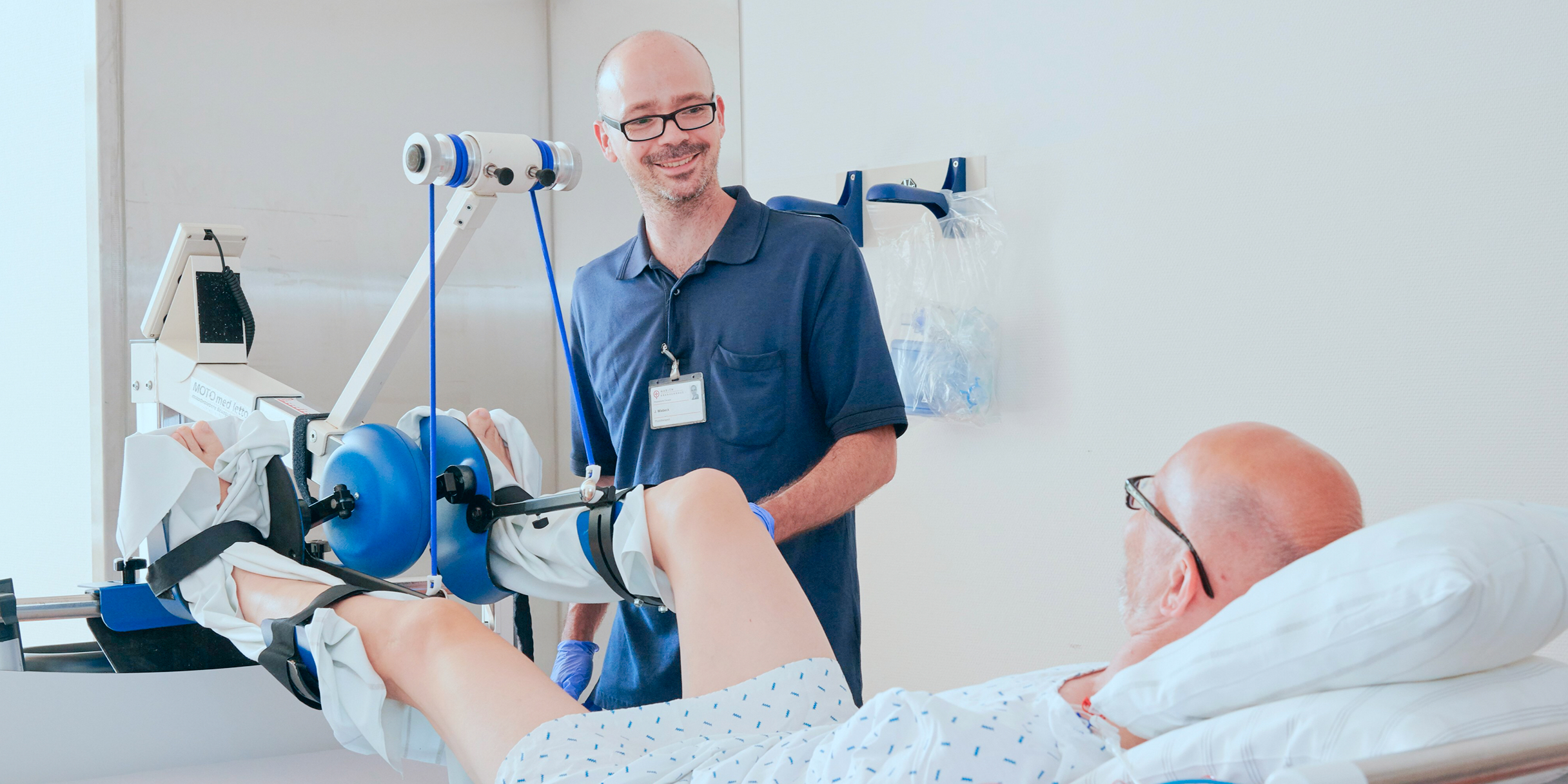 Ein Arzt steht neben einem Patienten im Krankenhausbett, der eine Rehabilitationsmaschine für das Bein benutzt.